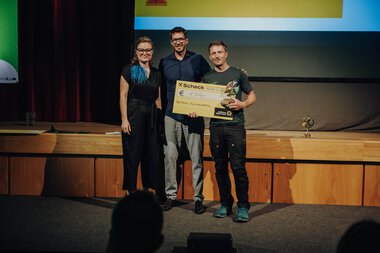 Drei Personen stehen auf einer Bühne, eine hält einen großen Scheck, bei einer Veranstaltung im Kleinwalsertal. Es ist eine Preisverleihung oder Anerkennung.