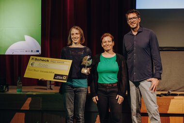 Drei Personen auf einer Bühne bei der Preisverleihung des Footprint Festivals 2024 im Kleinwalsertal. Die Frau auf der linken Seite hält eine Trophäe und ein großes Scheck.
