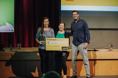 Drei Personen stehen auf einer Bühne, eine Frau hält einen großen Scheck und alle lachen, während sie an einer Veranstaltung teilnehmen.